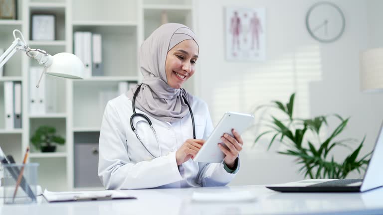 Muslim female doctor in a white coat is using browsing typing tablet in a modern hospital clinic.