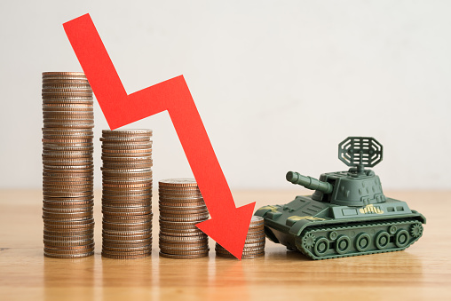 Economic crisis impact of Russian invasion of Ukraine concept. Stacked coins, graph falling down and battle tank on wooden table background copy space. War effect to world economy.