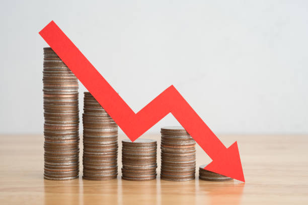 istock Stack of coins bar chart and red graph trending downwards with white wall background on wooden table. 1605302787