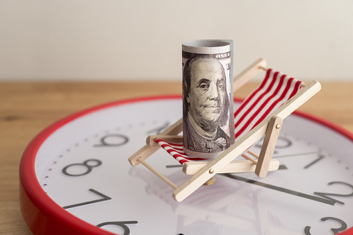 US dollar bill banknote on beach chair and red clock wooden table background. Concept of dividend yield in long term investment trading, passive income freedom, revenue from renting, pension from retirement.