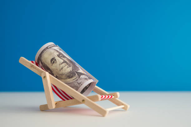 Happy US dollar bill banknote rest on wooden beach chair with blue background copy space in summer beach holiday. Financial freedom through passive income, success wealth from investment concept.