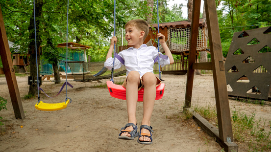 Happy laughing boy having fun while swinging on the playground at park.