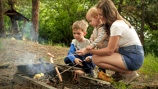 Young mother with two sons cooking sausages on bonfire in camp. Active leisure, children in camping, family vacation in nature