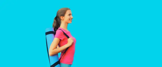 Sport and fitness concept - portrait of happy smiling young woman in sportswear with yoga mat looking away on blue background