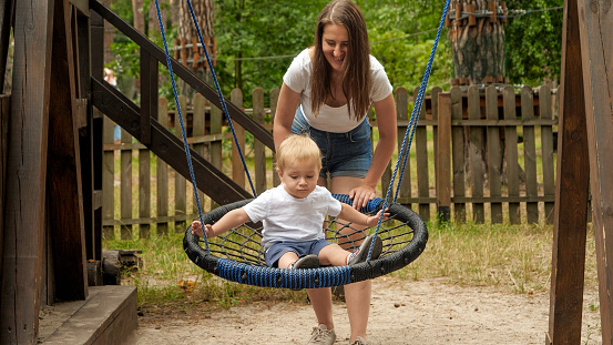 Young mother pushing her baby son swinging in rope nest swing in park. Kids playing outdoors, children having fun, summer vacation.
