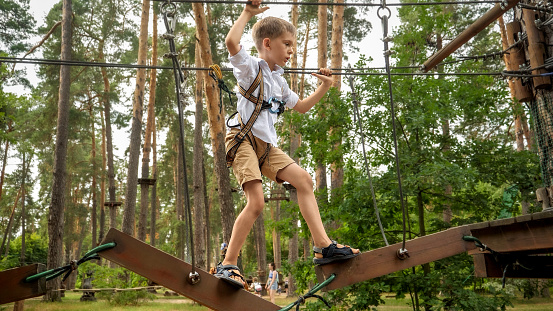 Young boy holding ropes and trying to cross the wobbly bridge in adventure park at forest.