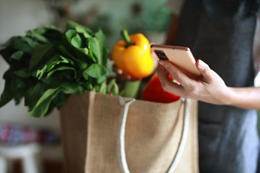 A woman's hand holds a smartphone over a table of fresh, colorful, and healthy fruits and vegetables from grocery shopping.. This scene portrays concepts of food delivery, responsible shopping, zero waste, sustainable lifestyle, and the intersection of lifestyle and technology.