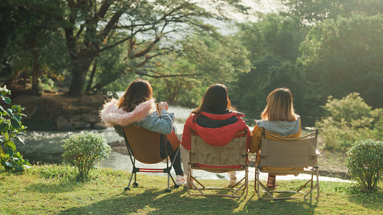 Group of happy Asian friends talking and drinking a cup of hot coffee for breakfast near stream in vacation with camping caravan at morning. Lifestyle travel nature concept.