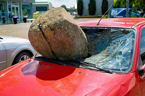 A large round stone rests on the hood of a red car with a shattered windshield.