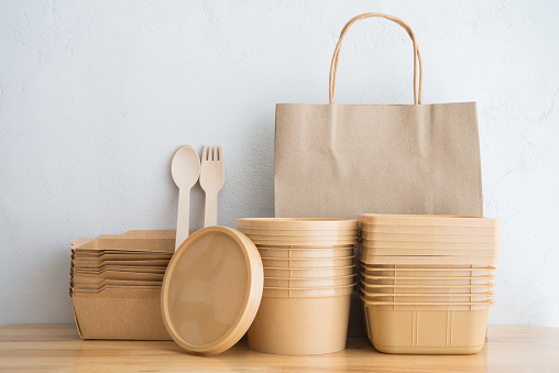 Natural eco-friendly disposable utensils (fork, spoon, paper bag, food box container, bowl) made of fiber of plant tree on wooden table with white wall background. Save the earth and waste reduction concept.