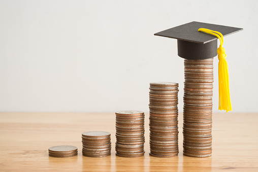 Education growth and development from scholarship, money saving or loan for education abroad concept. Coins stacked graph growing up and graduation hat on wooden table background copy space.