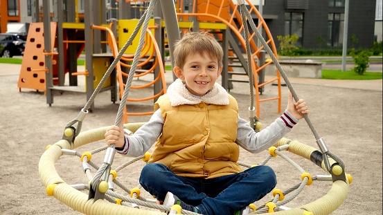 Happy smiling boy playing on public playground and spinning in net swing. Active child, sports and development, kids playing outdoors