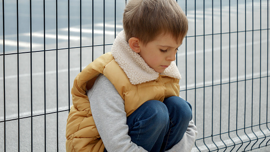Upset lonely boy being bullied at school sitting next to metal fence at playground. Child depression, problems with bullying, victim in school, emigration, criminal and poverty.