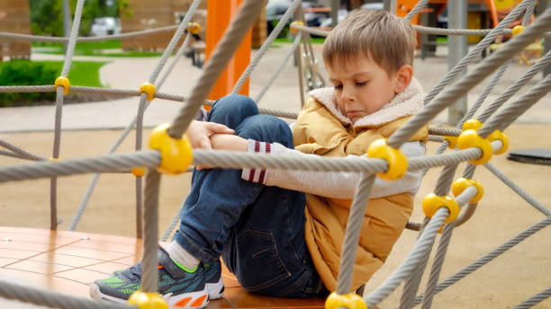 Upset lonely boy sitting on the playground and embracing his legs. Child depression, problems with bullying, victim in school, emigration, criminal and poverty. Upset lonely boy sitting on the playground and embracing his legs. Child depression, problems with bullying, victim in school, emigration, criminal and poverty sad teenager boy crying outdoors in park stock pictures, royalty-free photos & images