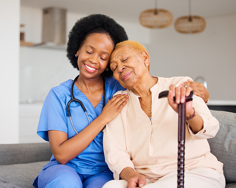 Black woman, nurse and senior patient in elderly care, hug and healthcare on living room sofa at home. Happy African medical professional or caregiver helping old age female person with cane in house