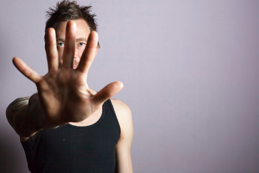portrait of a mid adult man wearing a black tank top looking through his hand which is held up and fingers are spread revealing his blue eyes behind.  horizontal composition with selective focus on background man's eyes.