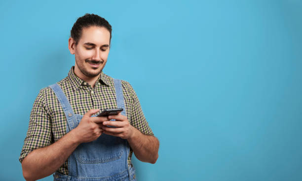 istock Technology savvy young male farmer with overalls smiling and texting with smartphone over blue background with copy space 1605434110