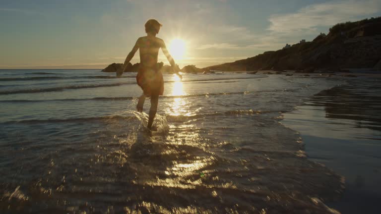 Boy run on the ocean beach splashing in water waves over sunset