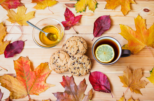 Cup of tea with lemon, honey in glass bowl, cookies with chocolate drops, and colorful autumn leaves on wooden table. Top view.