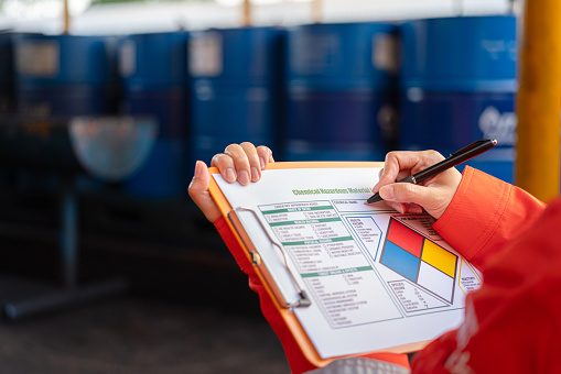 A safety office is checking on the hazardous material checklist form with chemical storage area at the factory as background. Industrial safety working scene, selective focus.
