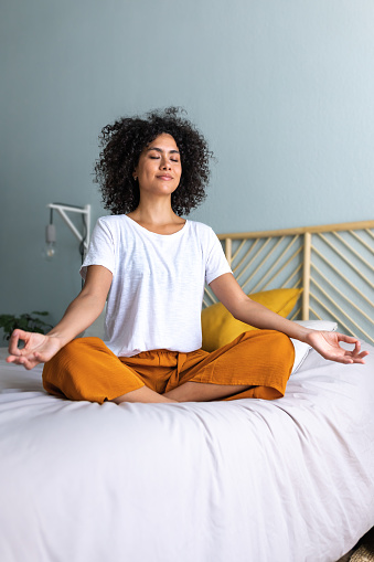 Young multiracial latina woman wearing comfortable clothes meditating on the bed to clear mind and relax. Mental health. Vertical image. Wellness and meditation concept.
