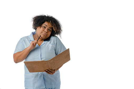 Afro female doctor wearing blue uniform looking at the camera talking on the phone and thinking resting the pen on her chin, while making notes, white background with copy space.