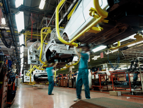 Two workers wearing green shirts and blue pants work underneath a car in an automobile factory.  There are assembly lines, partially constructed cars, tools and electrical equipment in the background.