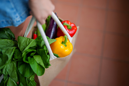 A cropped shot of a woman carrying an eco bag and shopping for groceries, holding a variety of fruits and vegetables. This scene portrays concepts of food delivery, responsible shopping, zero waste and sustainable lifestyle