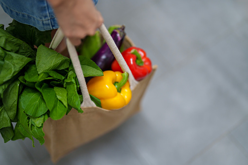 A cropped shot of a woman carrying an eco bag and shopping for groceries, holding a variety of fruits and vegetables. This scene portrays concepts of food delivery, responsible shopping, zero waste and sustainable lifestyle