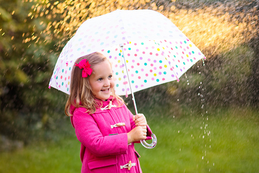 Little girl playing in rainy summer park. Child with colorful rainbow umbrella, pink coat walking in the rain. Kid having fun in autumn shower. Outdoor activity by any weather
