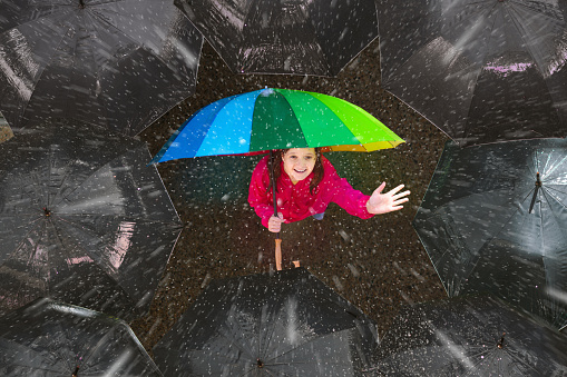 Happy young girl under colorful umbrella in dark crowd in autumn rain. Happiness and optimism concept. Joy and hope in difficult situations. Problem solving and creativity. Stand out and be unique.