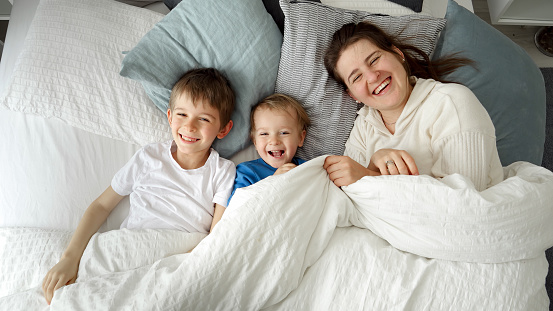 Happy mother with sons lying in bed under blanket and throwing it off smiling at camera. Concept of family happiness, relaxing at home, having fun in bed, parent and cheerful kids.