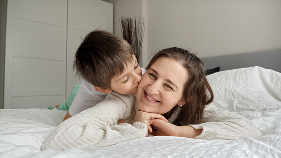 Portrait of cute boy lying in bed with mother and kissing her in cheek.