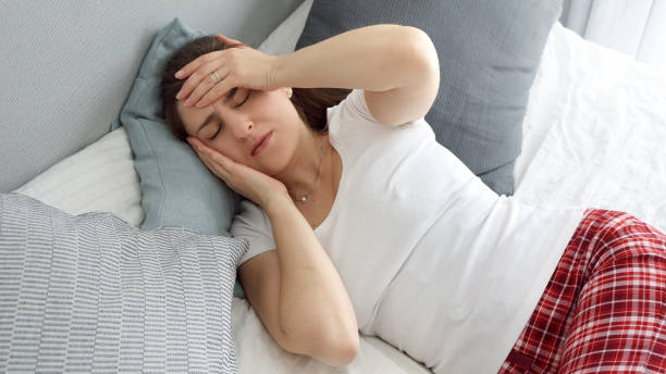 istock Young sick woman having high temperature lying in bed and holding hand on forehead while suffering from headache 1605615744