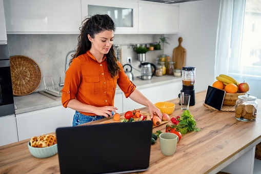 The woman is in the kitchen, she prepares a meal while using a laptop