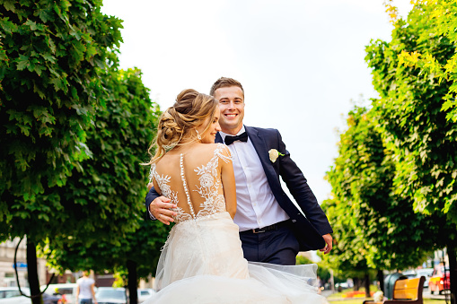The groom hugs his pretty bride in a chic dress with lace on the background of trees.