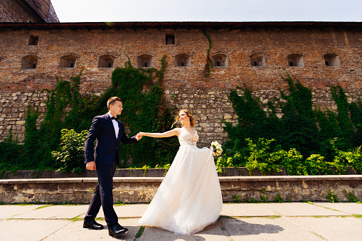 the bride holds wedding bouquet and leads the groom to the background of the fortress