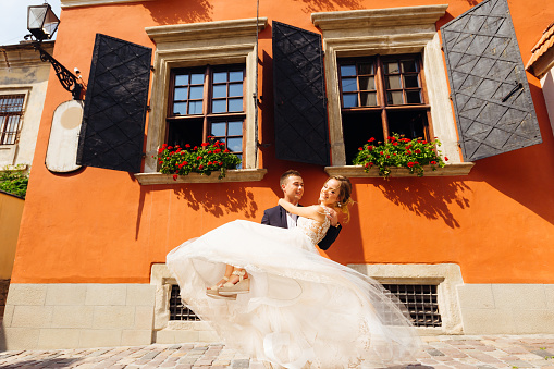 beautiful groom holds his pretty bride in his arms and they dance near the orange building on a beautiful sunny day