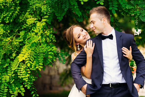 The bride embraces her groom in a beautiful suit and bow tie on the background of a green tree.