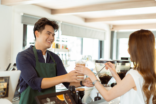 Asian male barista handing iced coffee to female customer over the counter at coffee shop cafe
