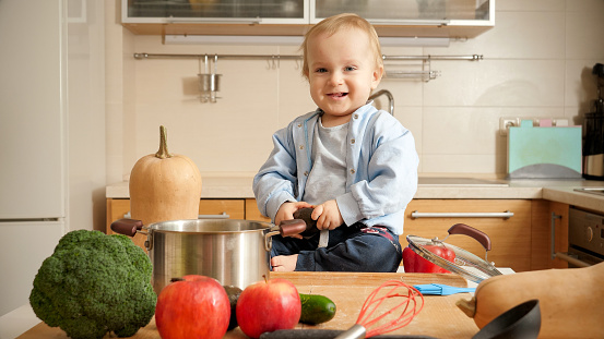 Portrait of happy smiling baby boy sitting on kitchen table and looking in camera. Concept of little chef, children cooking food, good family time together