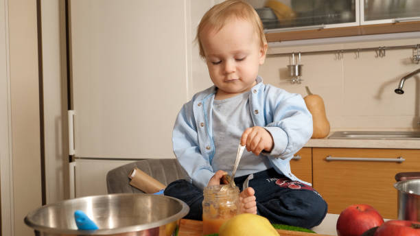 istock Little 1 year old baby boy sitting on kitchen table and eating fruit puree with spoon 1605744549
