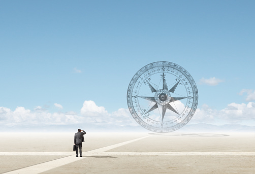A businessman stands at a crossroad as he places his hand to his forehead and looks into the disance at a compass that stands on the horizon.