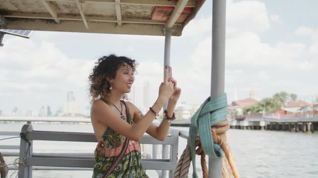 Cheerful travelers enjoying the scenery and taking pictures on their phones while riding a ferry on the Chao Phraya River in Bangkok.