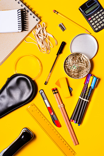 High angle view of school or office supplies over yellow color background as plastic transparent ruler,stapler, paper clips, pencil, exacto blade, rubber, sharpener, notebook, black magnifying glass case, calculator, color markers.