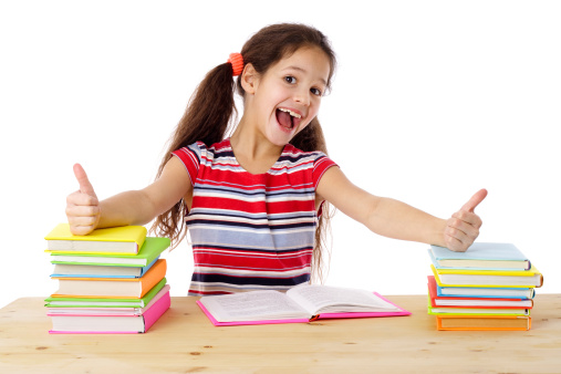 Girl with thumbs up sign sitting on the table with stack of books, isolated on white