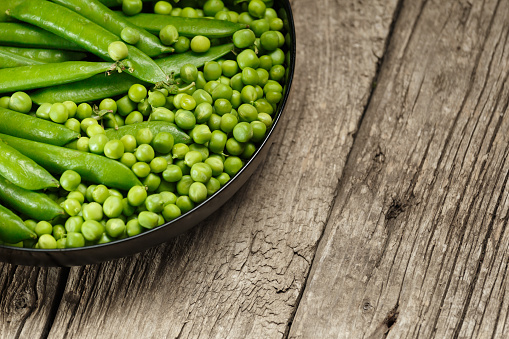 Green pea pods and shelled organic fresh peas in a round black plate on an aged wooden background, copy space. Vegetable protein, healthy products.