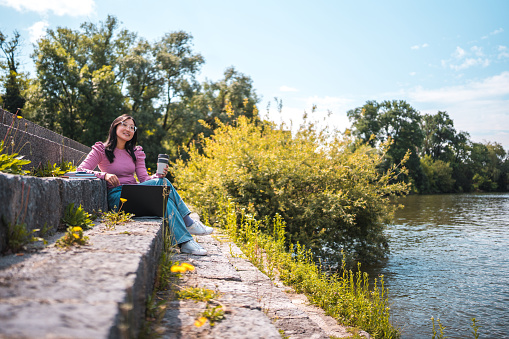 Full length image of a  young Asian female graduate with glasses, sitting on long, concrete stairs by a river and listening to music using a laptop. Wide angle shot. Take away coffee in her hand, trees in the background.