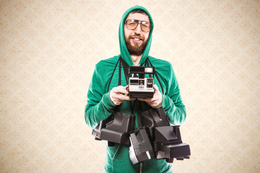 A bored young man with a beard, hooded sweatshirt, and vintage glasses has way too many Polaroid cameras hanging around his neck.  He looks at the camera makes a face like he thinks he's better than you.  Horizontal with retro wallpaper background.  Copy space available.