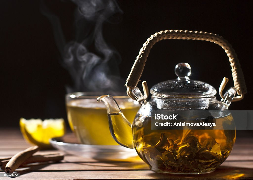 Glass teapot filled with tea sitting on a table with a cup  Green tea in Glass teapot  on wooden table .On the background steaming cup of tea,  piece of lemon and cinnamon sticks Black Background Stock Photo